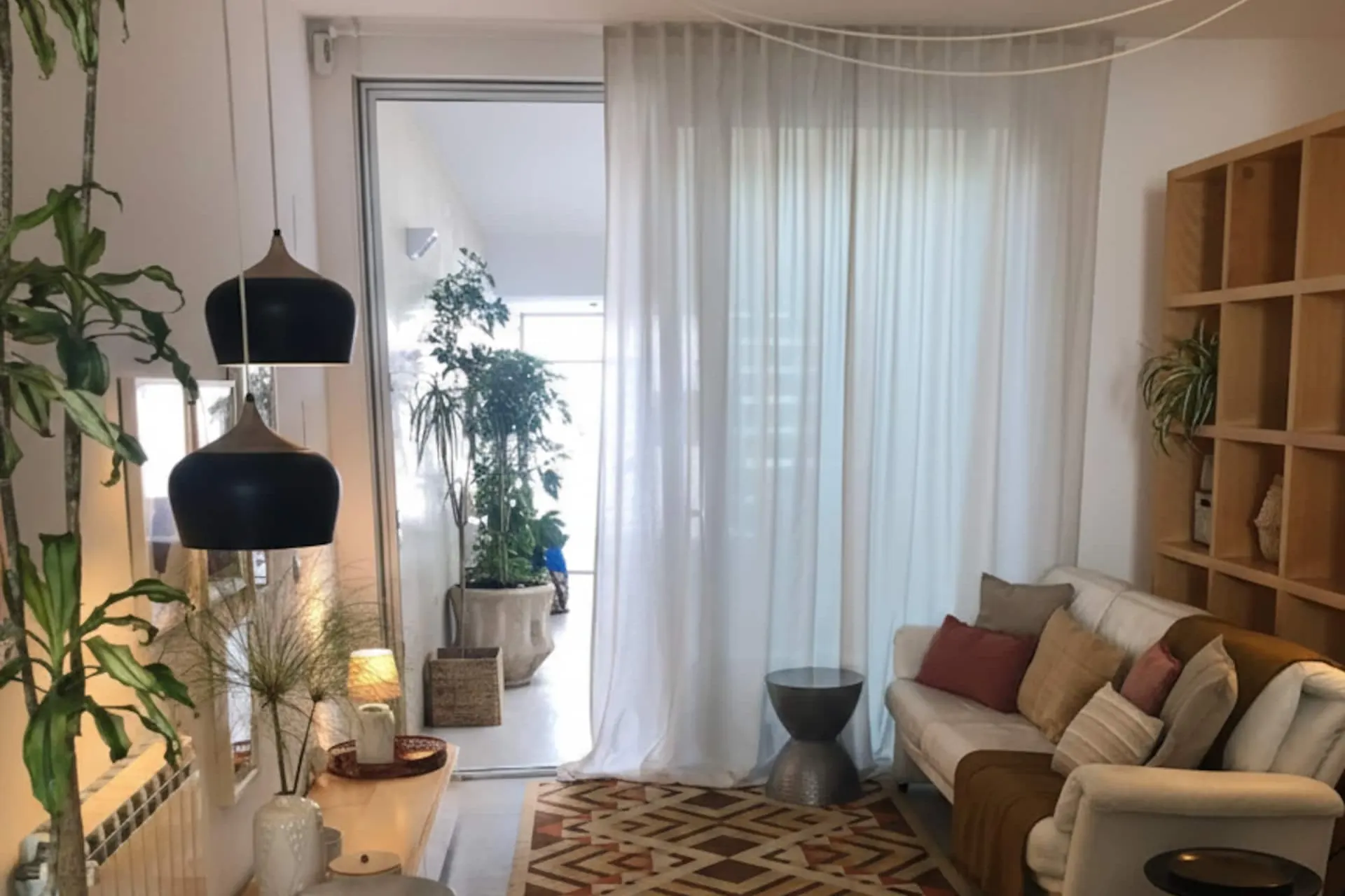 Sala em open space com sofá claro, tapete geométrico e cortinados brancos que abrem para o pátio